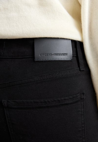 Mustmustest denim püksid, millel on nahast brändi silt, millel on reljeefne tekst. Nähtav õmblus ja tagakoti detail. Heledat värvi särk on osaliselt nähtav.