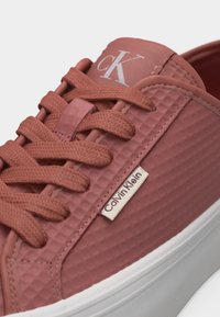 Gros plan sur une basket Calvin Klein rose texturée avec des lacets assortis, une étiquette logo blanche sur le côté et une semelle blanche.