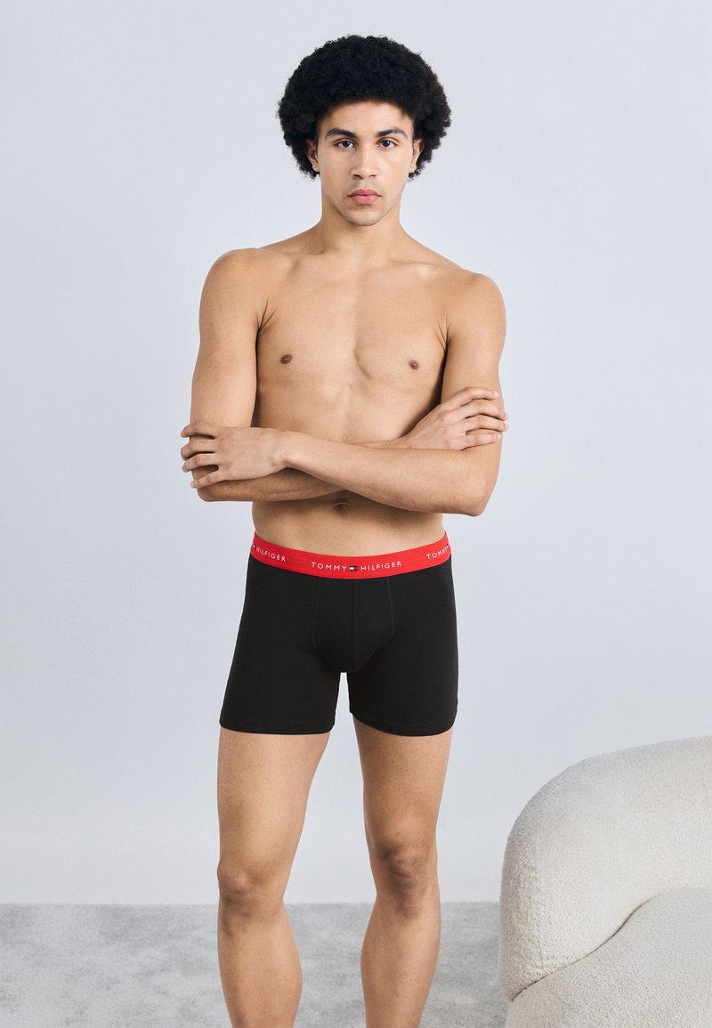 Μαύρα ανδρικά εσώρουχα boxer με κόκκινη μέση που φέρει το λογότυπο "TOMMY HILFIGER". Το ύφασμα φαίνεται λείο και η εφαρμογή είναι στενή στο σώμα.