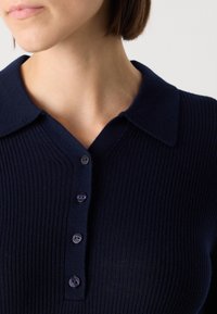 Polo shirt en tricot bleu marine avec une texture côtelée, doté d'un col doux et de cinq boutons violets le long de la patte de boutonnage.