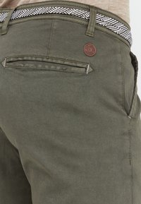 Pantalon vert olive avec une taille texturée présentant un motif noir et blanc. Comprend une petite étiquette en cuir marron et des poches arrière.
