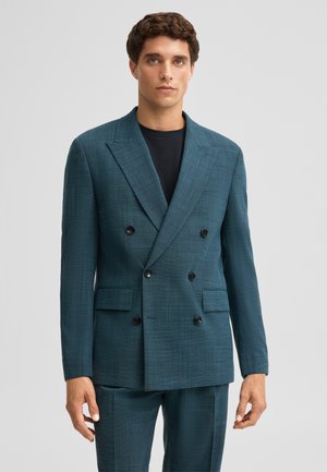 Man draagt een teal double-breasted pak met zwarte knopen en bijpassende broek over een zwart shirt met ronde hals, staand tegen een effen achtergrond.