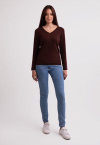 Brauner V-Ausschnitt-Pullover mit langen Ärmeln, kombiniert mit hellblauen, figurbetonten Jeans und weißen Sneakers. Glatte Textur, lässiges Design.