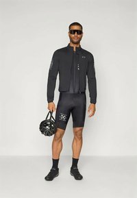 Mand iført sort cykeltøj og solbriller, der holder en sort cykelhjelm, stående mod en ensfarvet lys baggrund.