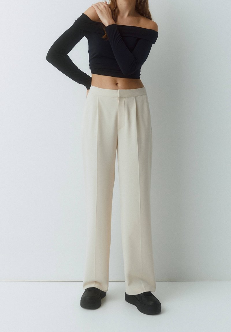 Black off-the-shoulder top paired with high-waisted, cream-colored, wide-leg trousers. Trousers feature front pleats and a smooth texture.