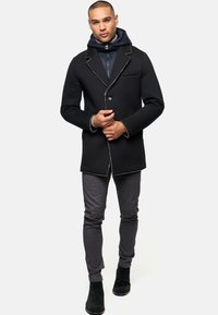 Black coat with contrasting white stitching, single button closure, and notch lapels, layered over a dark hooded jacket and gray pants.