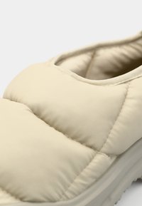 Chaussure slip-on beige avec une texture matelassée. Forme arrondie, semelle épaisse et col élastique. Matériau doux avec des détails de couture subtils.