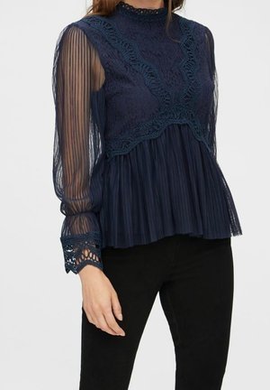 Kvinde iført en marineblå blondebluse med gennemsigtige, plisserede ærmer og bølgede manchetter, kombineret med sorte bukser.