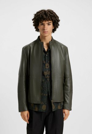 Olive green leather jacket with a stand collar, zippered front, and side pockets, worn over a patterned dark shirt. Smooth texture, tailored fit.