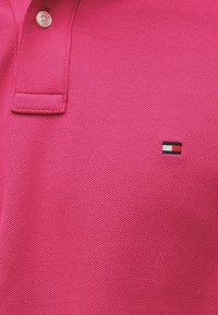 Πόλο πουκάμισο σε έντονο ροζ με υφασμάτινη υφή, διαθέτει κούμπωμα με τρία κουμπιά και μικρό κεντημένο λογότυπο σε κόκκινο, άσπρο και μπλε.