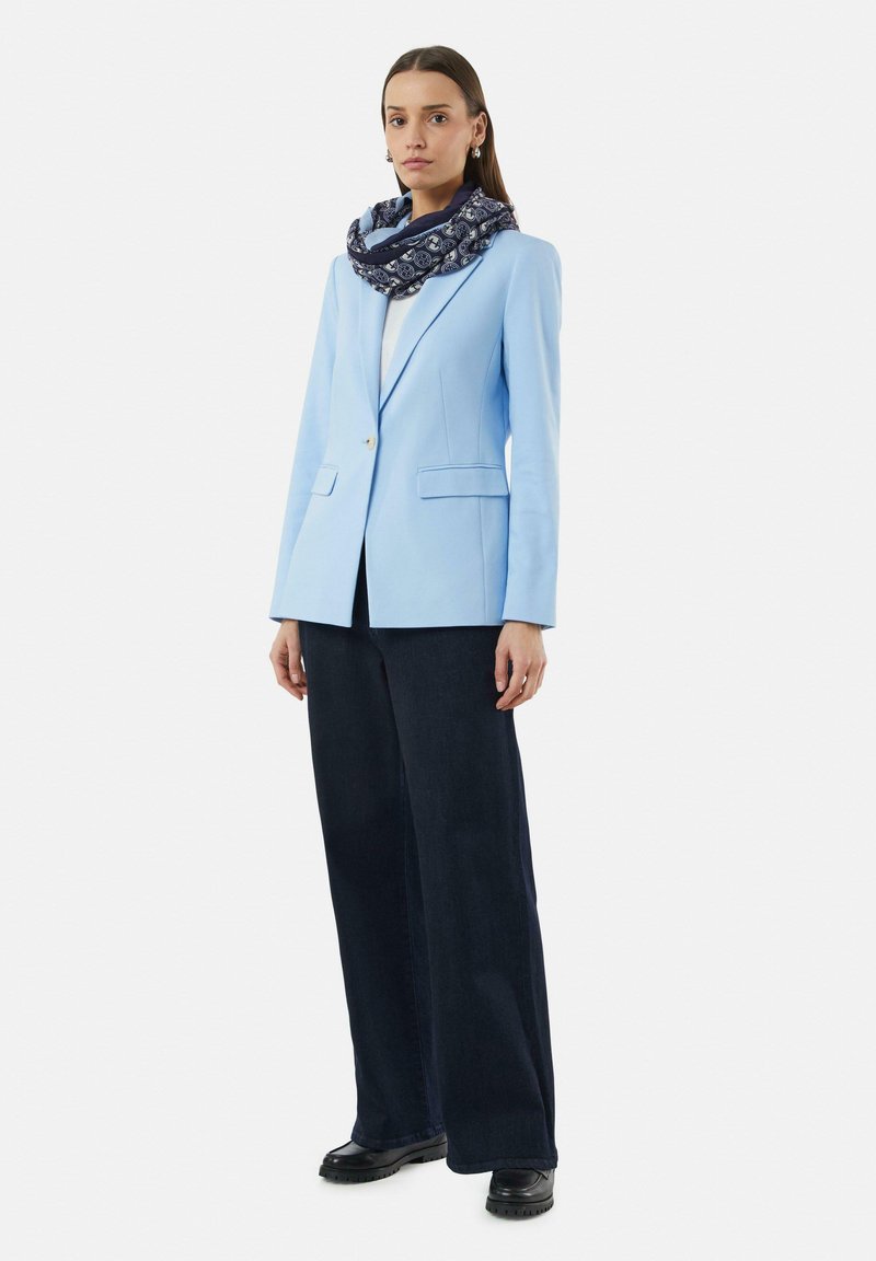 Light blue tailored blazer with two front pockets, paired with dark wide-leg jeans. A patterned navy scarf accentuates the look. Black footwear.