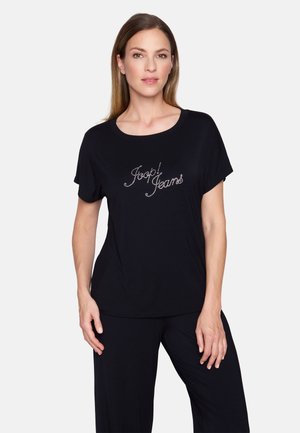 Femme aux cheveux bruns et lisses portant un haut noir à manches courtes avec un logo en strass "Joop! Jeans", debout avec une main sur la hanche.