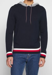 Pull en maille côtelée marine avec une capuche grise, une taille rayée rouge et blanche, et des cordons de serrage tressés. Il possède des manches longues et une coupe décontractée.