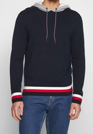 Pull en maille côtelée marine avec une capuche grise, une taille rayée rouge et blanche, et des cordons de serrage tressés. Il possède des manches longues et une coupe décontractée.