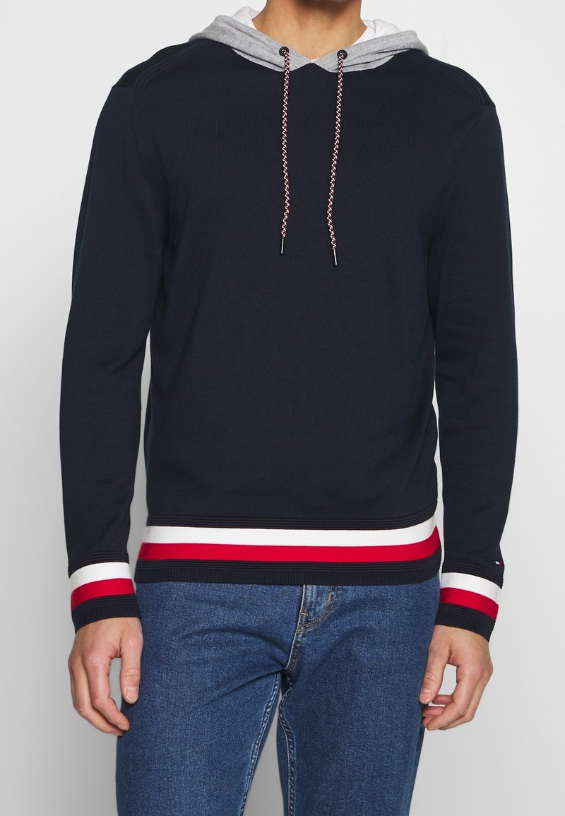 Pull en maille côtelée marine avec une capuche grise, une taille rayée rouge et blanche, et des cordons de serrage tressés. Il possède des manches longues et une coupe décontractée.