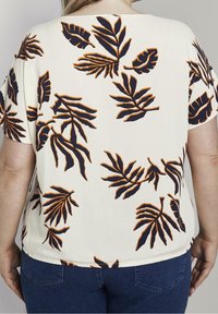 Top corto bianco a maniche corte con una grande stampa di foglie blu navy e arancioni, texture liscia e vestibilità rilassata, con orlo posteriore arrotondato.