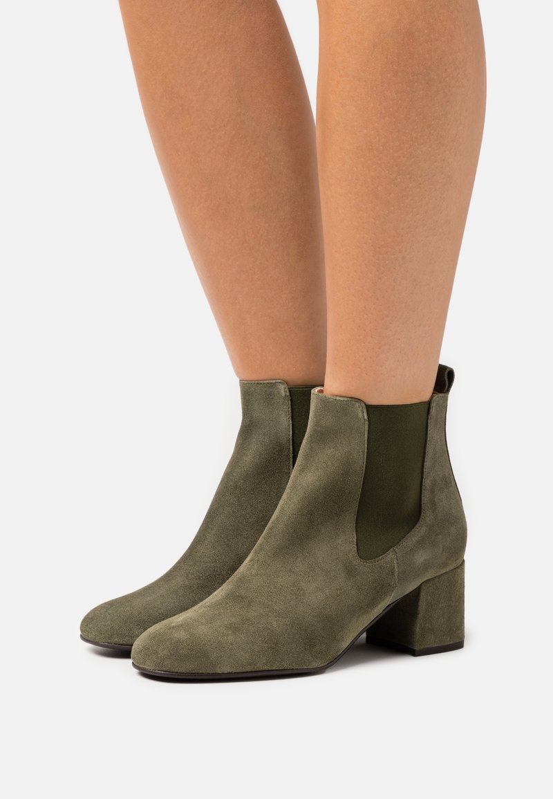 Paire de bottines en daim vert olive à talons blocs portées sur des jambes nues sur un fond clair uni.
