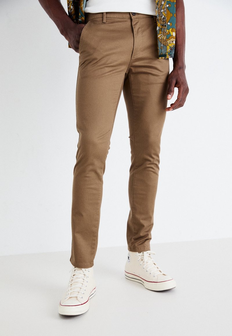 Hnědé slim-fit kalhoty vyrobené z bavlněné směsi. Mají klasický design kapes a jsou kombinovány s bílými vysokými teniskami.