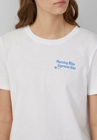 T-shirt en coton blanc à col rond, avec le texte bleu "Morning Bliss Espresso Kiss" sur le côté gauche de la poitrine. Texture lisse.