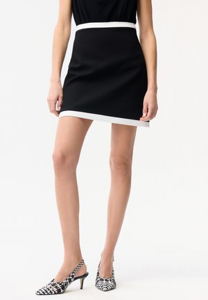 Vrouw draagt een zwarte minirok met witte bies en zwart-witte slingback hakken met patroon, staand tegen een effen witte achtergrond.