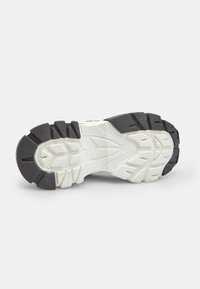 Suela de un zapato deportivo blanco y negro con profundos patrones de banda de rodadura para tracción, ubicado contra un fondo gris liso.