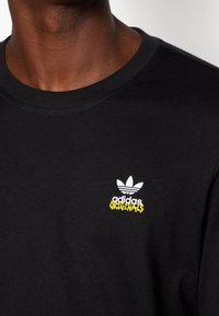 Svart bomullströja med rund hals, med en vit Adidas-logotyp och gul "ORIGINALS" text broderad på vänster bröstområde.