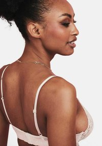 Lace bralette in een zachte beige kleur met verstelbare bandjes, een haak-en-oogsluiting en een bloemenmotiefaccent. Zijaanzicht dat het ontwerp aan de achterkant toont.