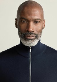 Mørkeblå zip-up trøje med høj krave, prydet med en glat tekstur og sølvfarvet lynlåsdetalje. Nærbillede af en mand.