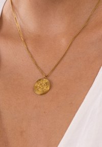 Goldfarbener Halsreifen mit einem ovalen Anhänger, der kunstvoll graviert ist und an einer dünnen Kette hängt. Der Anhänger hat eine strukturierte Oberfläche.