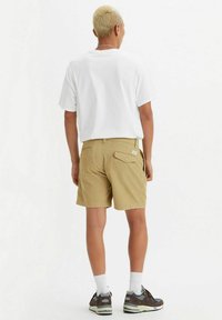 Shorts bej cu buzunar pe spate, asortați cu un tricou alb și pantofi sport negru-maro. Modelul are părul scurt și blond și șosete albe.
