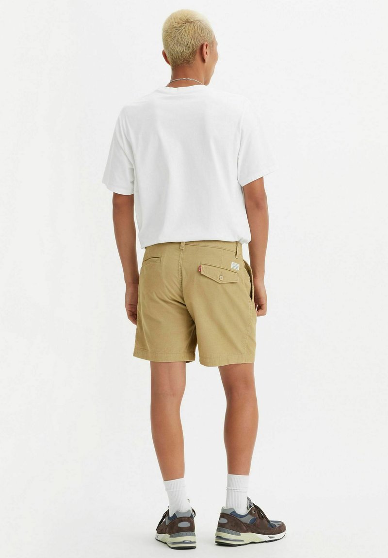 Pantalones cortos beige con un bolsillo trasero, combinados con una camiseta blanca y zapatos deportivos negro-marrones. El modelo tiene el cabello corto y rubio y lleva calcetines blancos.