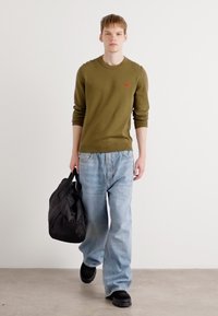 Pull vert olive avec logo rouge, associé à un jean bleu clair à coupe large et des chaussures noires à enfiler. Le mannequin porte un sac noir.