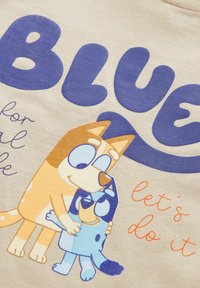 T-shirt lavet af blødt, lysfarvet stof med et legende grafisk motiv af to tegnefilmsagtige hunde, der krammer. Teksten inkluderer "BLUE" og "lad os gøre det."