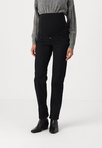 Persona embarazada vestida con jeans de maternidad negros, suéter de punto gris y botines negros, de pie contra un fondo blanco liso.