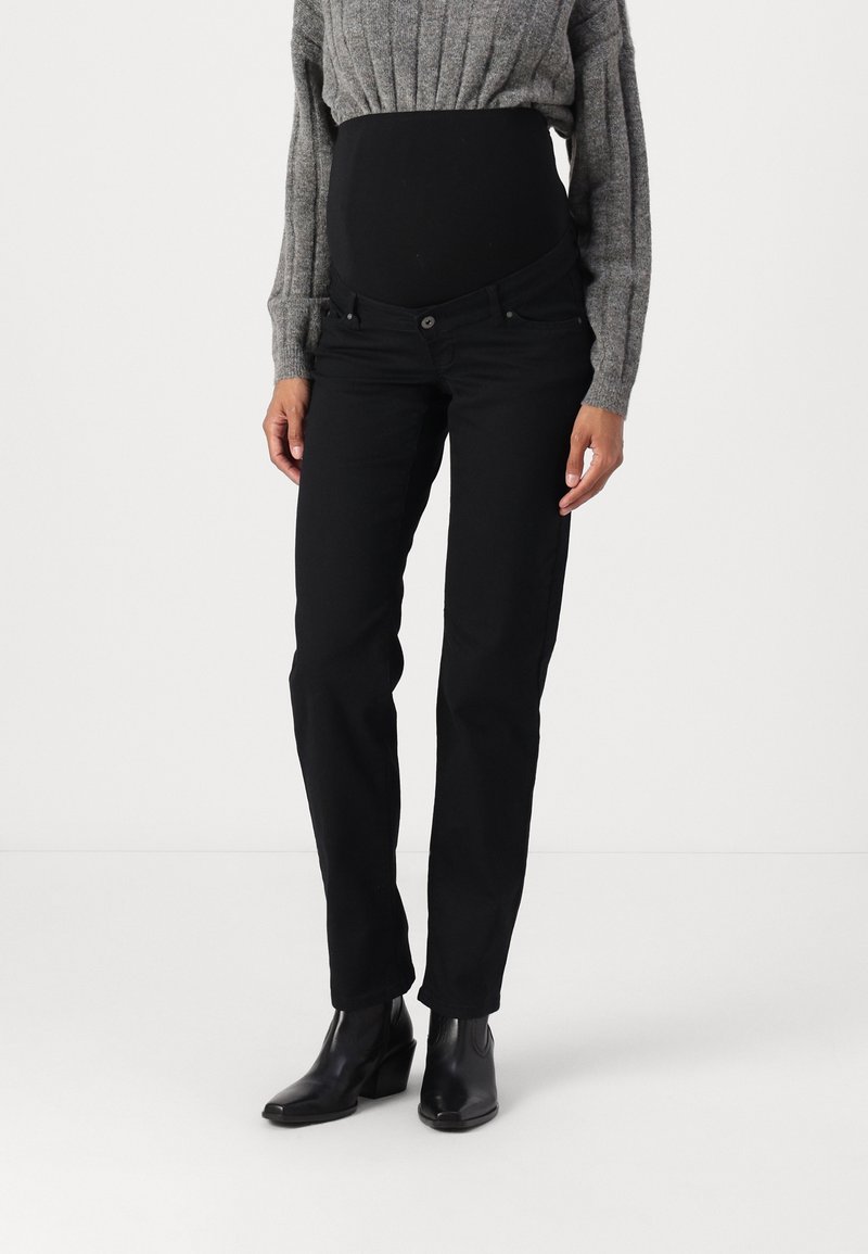 Persona embarazada vestida con jeans de maternidad negros, suéter de punto gris y botines negros, de pie contra un fondo blanco liso.