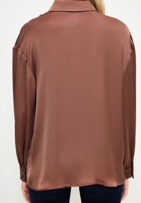 Langærmet, brun satin skjorte med krave. Har knappede manchetter og en glat tekstur, stylet med en forlænget ryg.