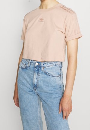 Femme portant un t-shirt Adidas cropped rose clair avec manches retroussées et un jean taille haute bleu clair, sur un fond uni.