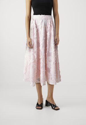 Femme portant une jupe midi en dentelle florale rose, un haut noir sans manches et des sandales noires à talon bloc ouvertes sur un fond uni.