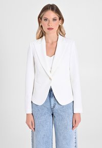Blazer blanc ajusté avec revers en pic, fermeture à un bouton et deux poches avant. Porté sur un haut crème et assorti à un jean bleu clair.