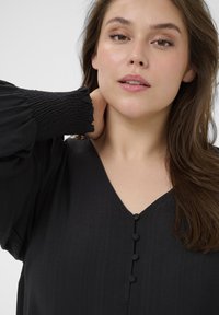 Sort bluse med V-udskæring, teksturerede puffærmer og tre dekorative knapper foran. Glat stof med subtil glans.