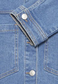 Jachetă din denim albastru deschis văzută în plan apropiat, care prezintă manșetă pliată, nasturi metalici cu capse și buzunare cusute vizibil cu ață portocaliu-maro.