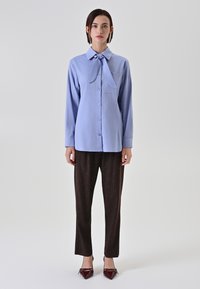 Camicia azzurro chiaro a maniche lunghe con colletto a farfallino, abbinata a pantaloni scuri a fantasia marrone e scarpe bordeaux a punta.