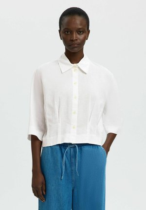 Naine kannab valget poolvarrukate ja nööpidega särki ning lahtisi siniseid tõmblukuga pükse, seisab käsi taskus.