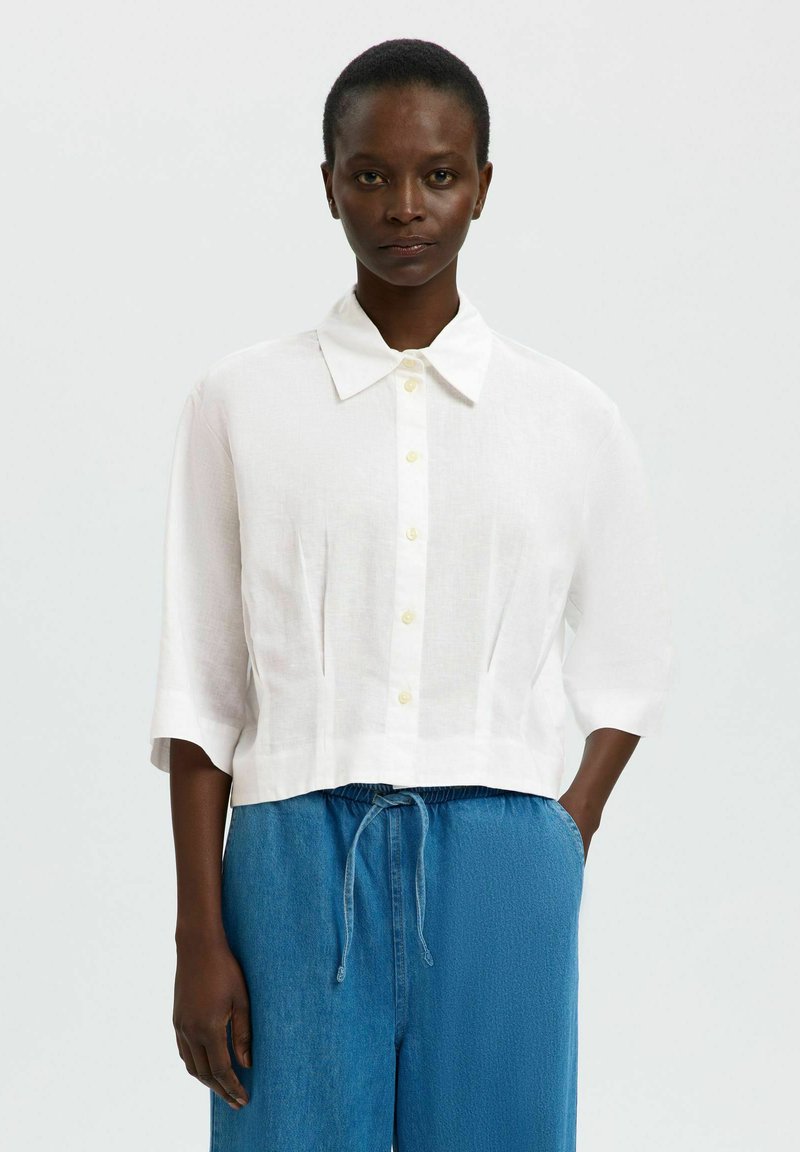 Femme portant une chemise blanche boutonnée à manches mi-longues et un pantalon bleu ample avec cordon, debout avec une main dans la poche.