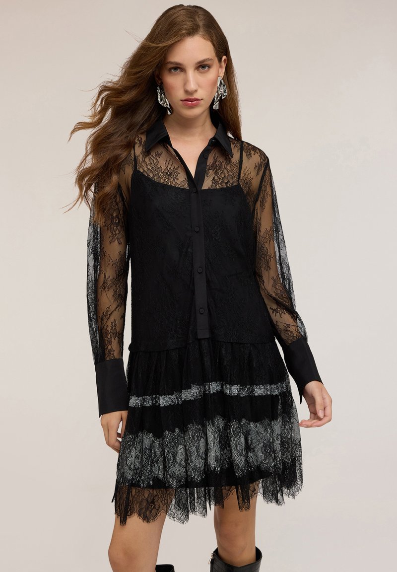 Schwarzes Spitzenkleid mit durchsichtigen langen Ärmeln, vorne mit Knöpfen und einem geschichteten Rock mit silbernen Akzenten und strukturierten Details.