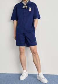 Mörkblå kortärmad skjorta och matchande shorts, tillverkade av ett slätt tyg med en liten broderad blomstertid. Kombinerat med vita sneakers.