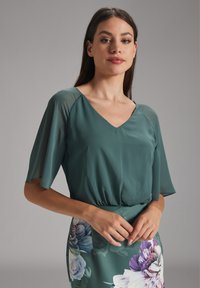 Blouse turquoise avec un décolleté en V et des manches flottantes, assortie à une jupe à motifs floraux comportant de grandes fleurs violettes et blanches.