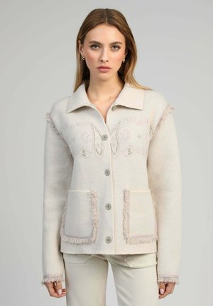 Femme portant une veste en laine beige clair boutonnée avec des franges sur les poches et les poignets, debout devant un fond gris uni.