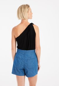 Top negro de un solo hombro con detalle de lazada, combinado con pantalones cortos de mezclilla azul de cintura alta y bolsillos traseros. Textura suave y ajuste relajado.