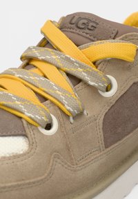 Gros plan d'une basket beige UGG avec des lacets jaunes et gris à motifs, des panneaux en suède cousus et des œillets blancs sur une semelle claire.
